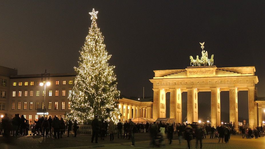 Christmas tree lit up in front of the illuminated Brandenburg Gate in Berlin at night, with crowds of people.