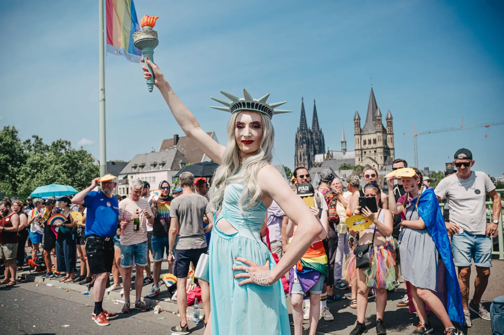 A person dressed as the Statue of Liberty poses at a Pride parade in front of the Cologne Cathedral on a sunny day.