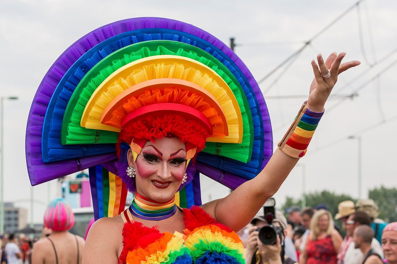 A person in a large, rainbow-colored fan headdress and matching outfit waves at an outdoor pride parade.