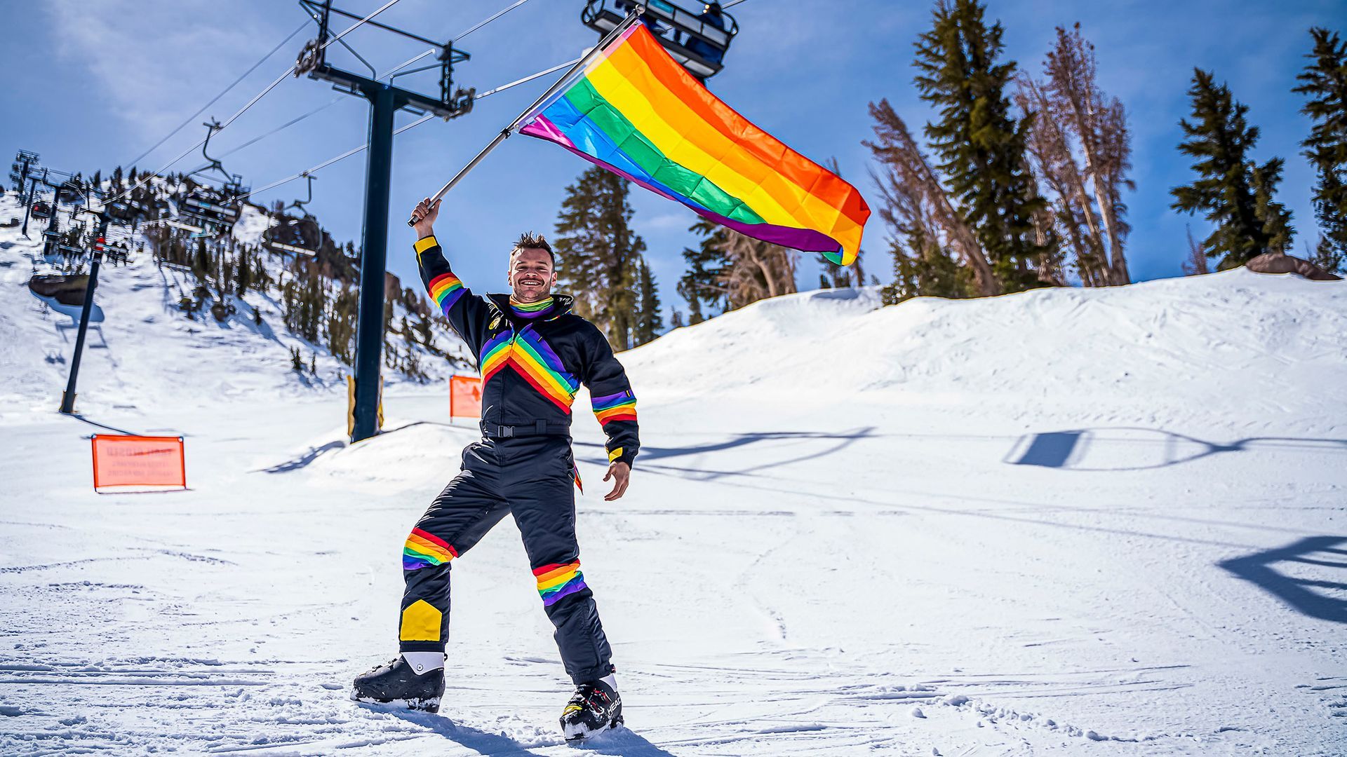 Person in rainbow ski suit holding a rainbow flag on a snowy mountain with ski lift.