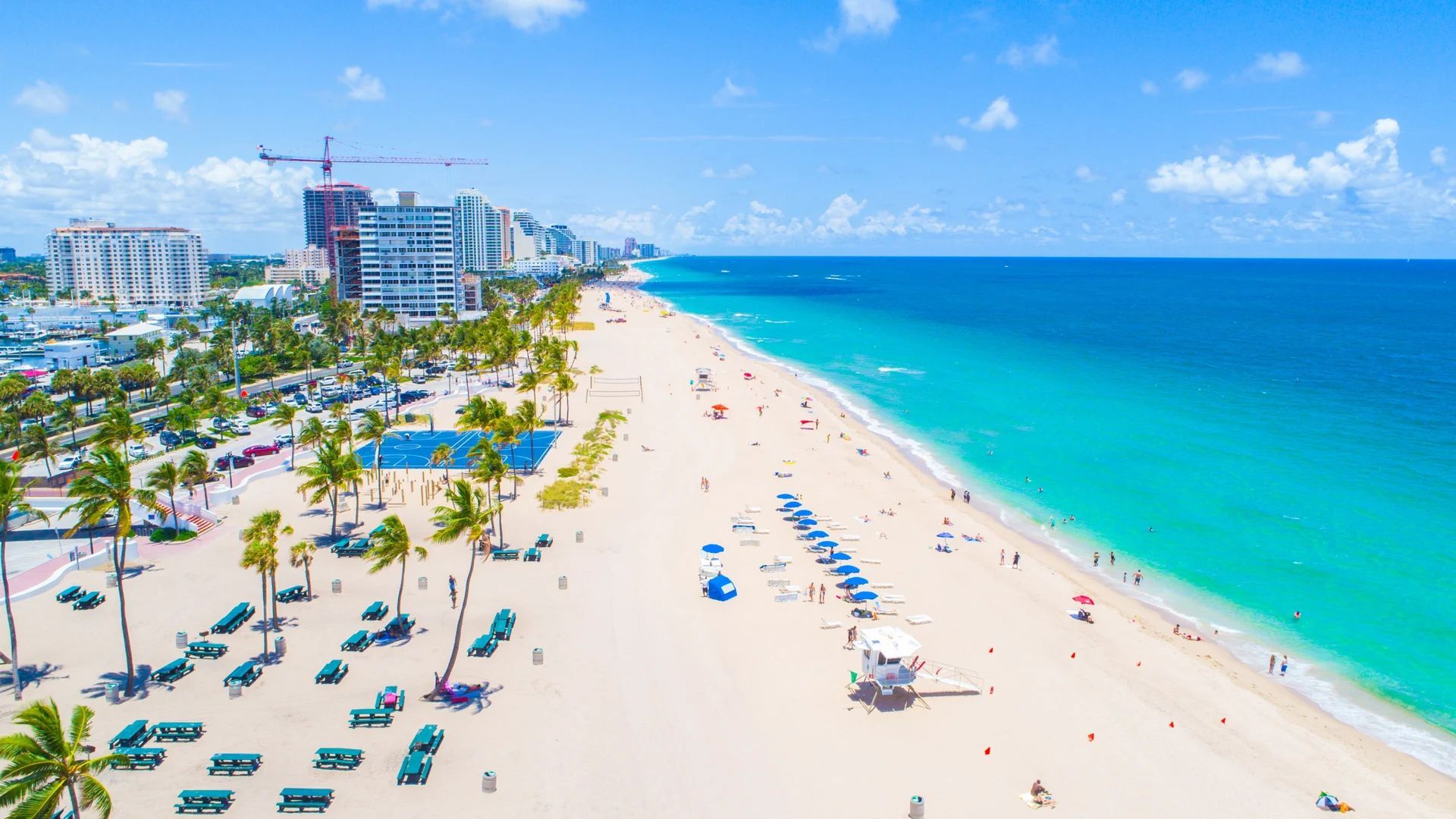 Aerial view of a beach with white sand, turquoise water, palm trees, and hotels under a blue sky. Fort Lauderdale