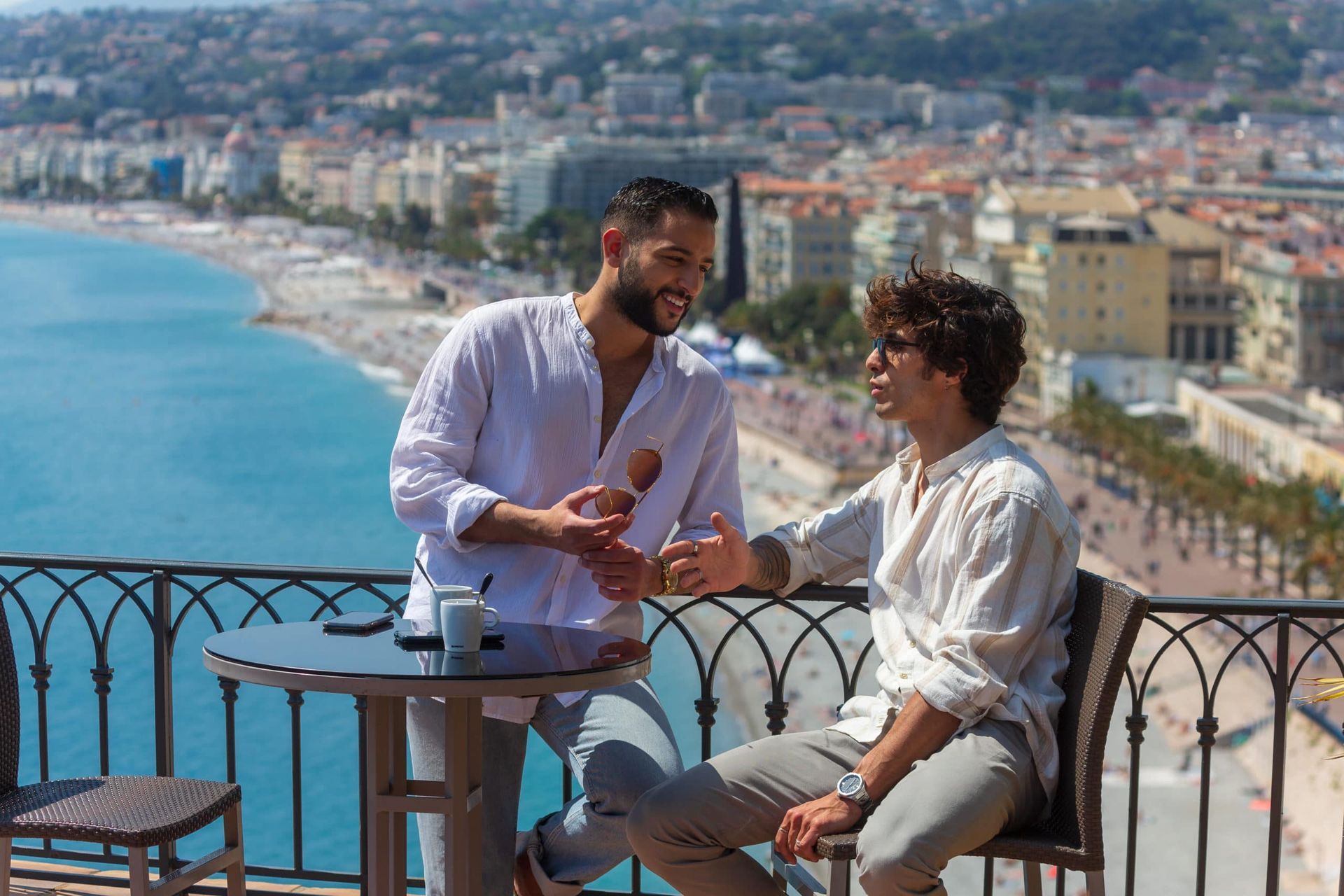 Two men on a balcony overlooking a beach. One holds an ice cream cone; both are smiling and talking. Bright sunny day.