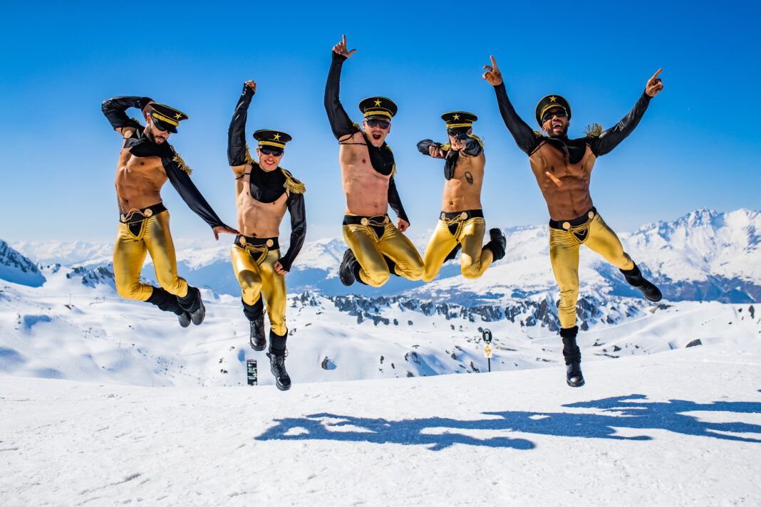 Five shirtless people in gold pants and hats jump on a snowy mountain, arms raised.