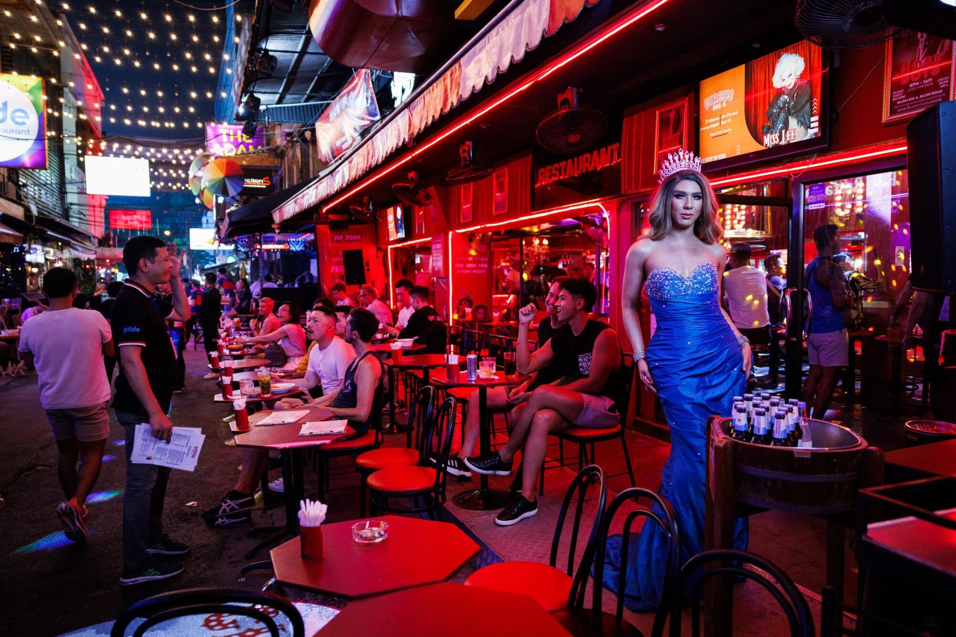 Night street scene with neon lights, outdoor tables, and a woman in a blue gown posing.