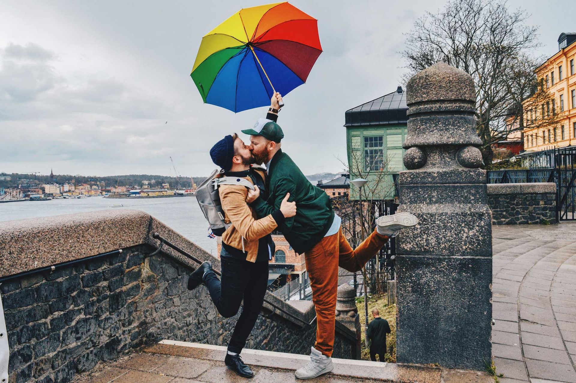 Two people kissing under a rainbow umbrella, outdoors near water. One person kicks up a leg.