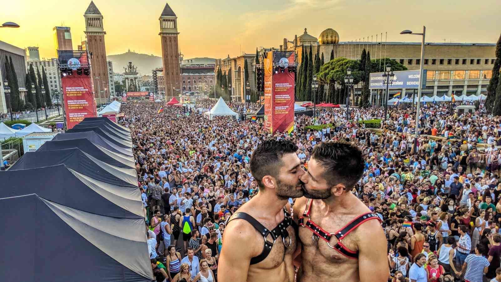 Two men wearing leather harnesses kissing during a crowded outdoor festival at sunset, with a city plaza in the background.