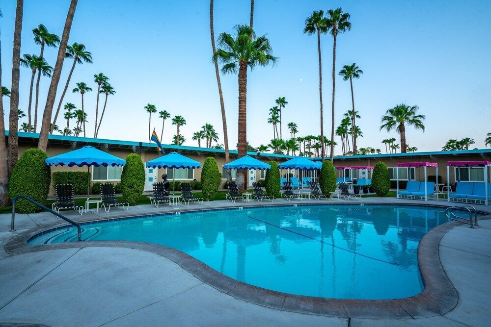 A turquoise swimming pool surrounded by palm trees, lounge chairs, and blue umbrellas against a clear sky.