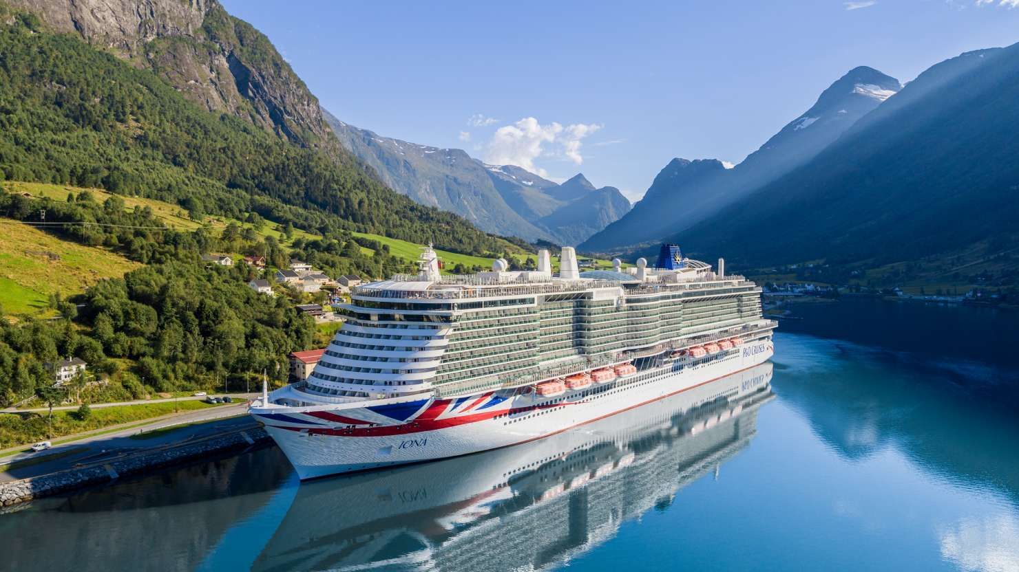 Cruise ship docked in a fjord, mountains in the background, blue water and sky.