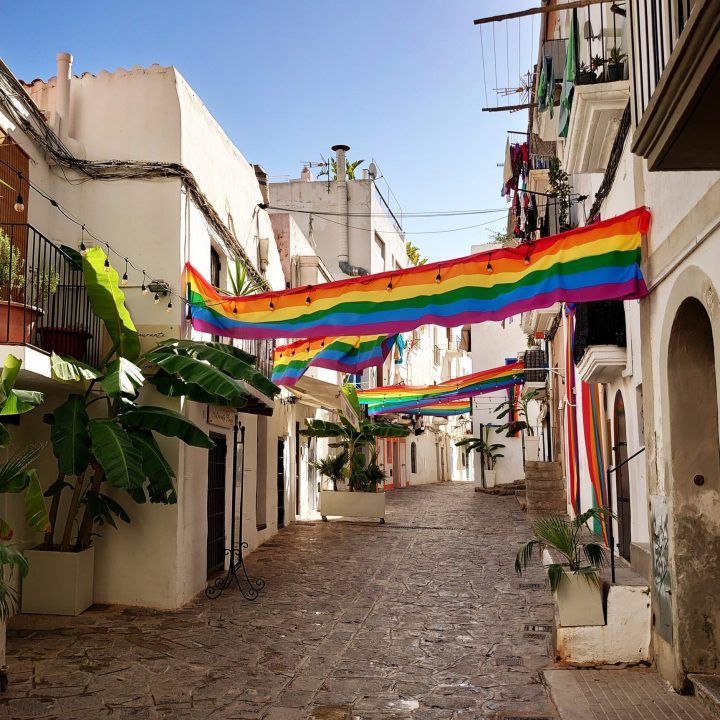Narrow cobblestone street lined with white buildings, adorned with rainbow flags. Ibiza
