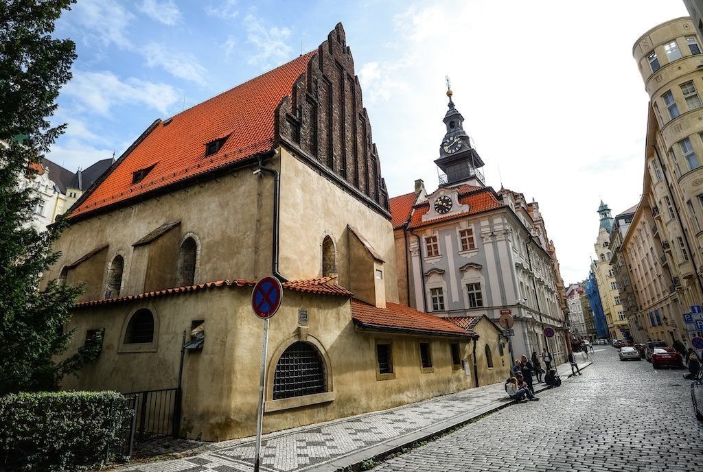 Old Synagogue in Prague with red-tiled roof and tall, ornate clock tower in the background on a cobblestone street. Josefov
