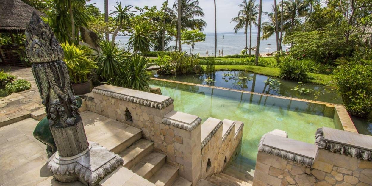 Stone steps lead to a pool overlooking a beach with palm trees.
