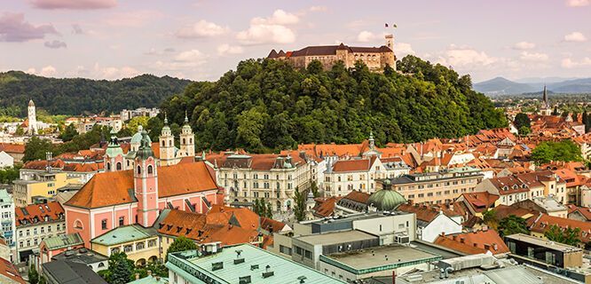 A scenic view of Ljubljana Castle atop a lush green hill, overlooking a historic city with red-roofed buildings. Ljubljana