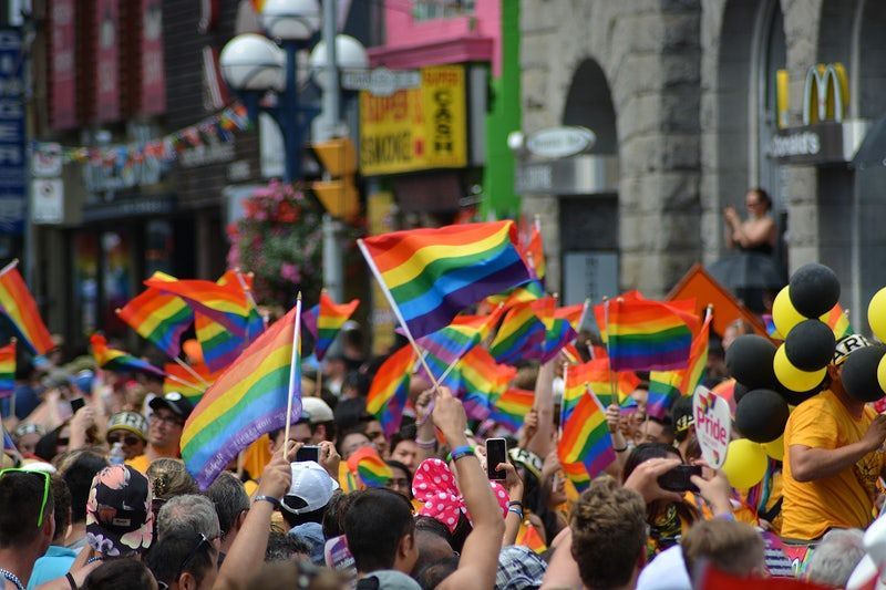 A crowd celebrates at a pride parade, waving numerous rainbow flags while holding yellow and black balloons.
