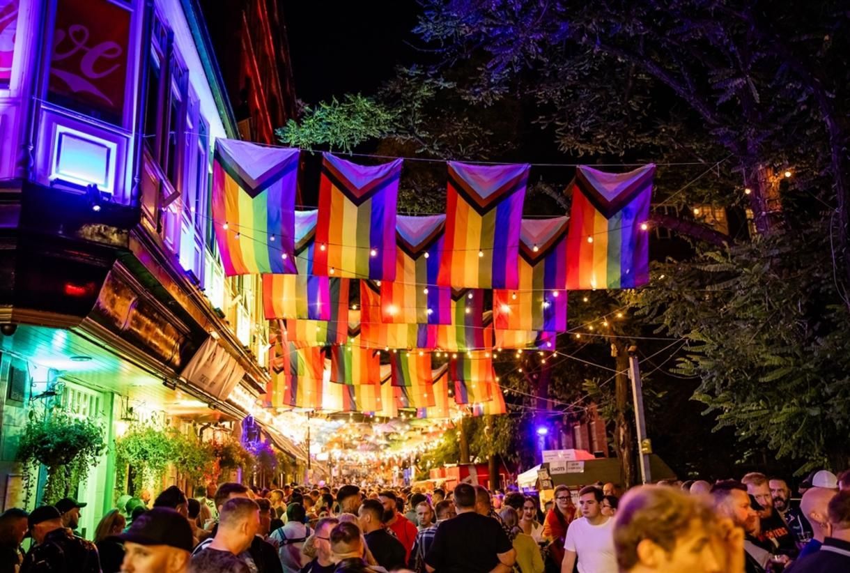 Crowded nighttime street festival with rainbow flags and colorful lights hanging overhead