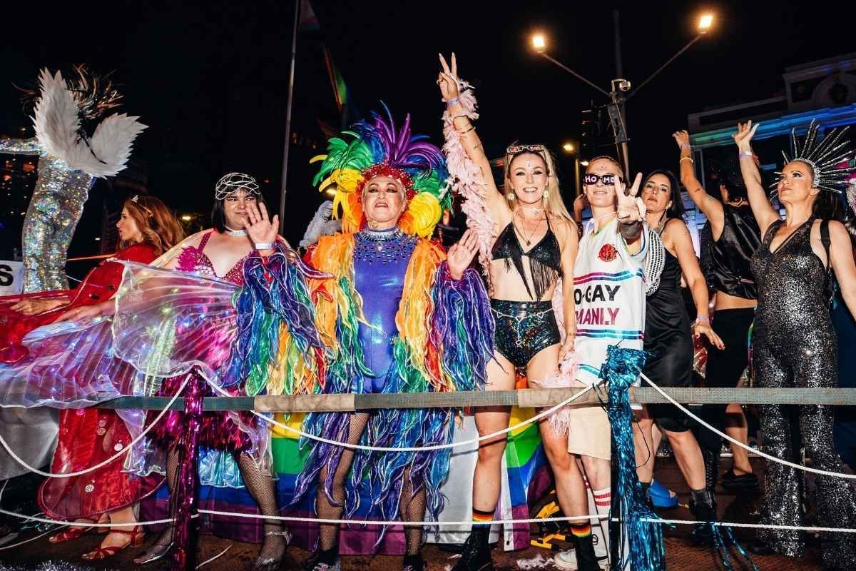 A group of people in festive, colorful costumes and glittery outfits pose on a parade float at night. Sydney Mardi Gras