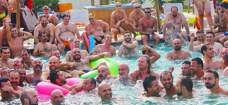 A large group of men in a swimming pool, many are bare-chested. Several have facial hair and floaties. gran canaria