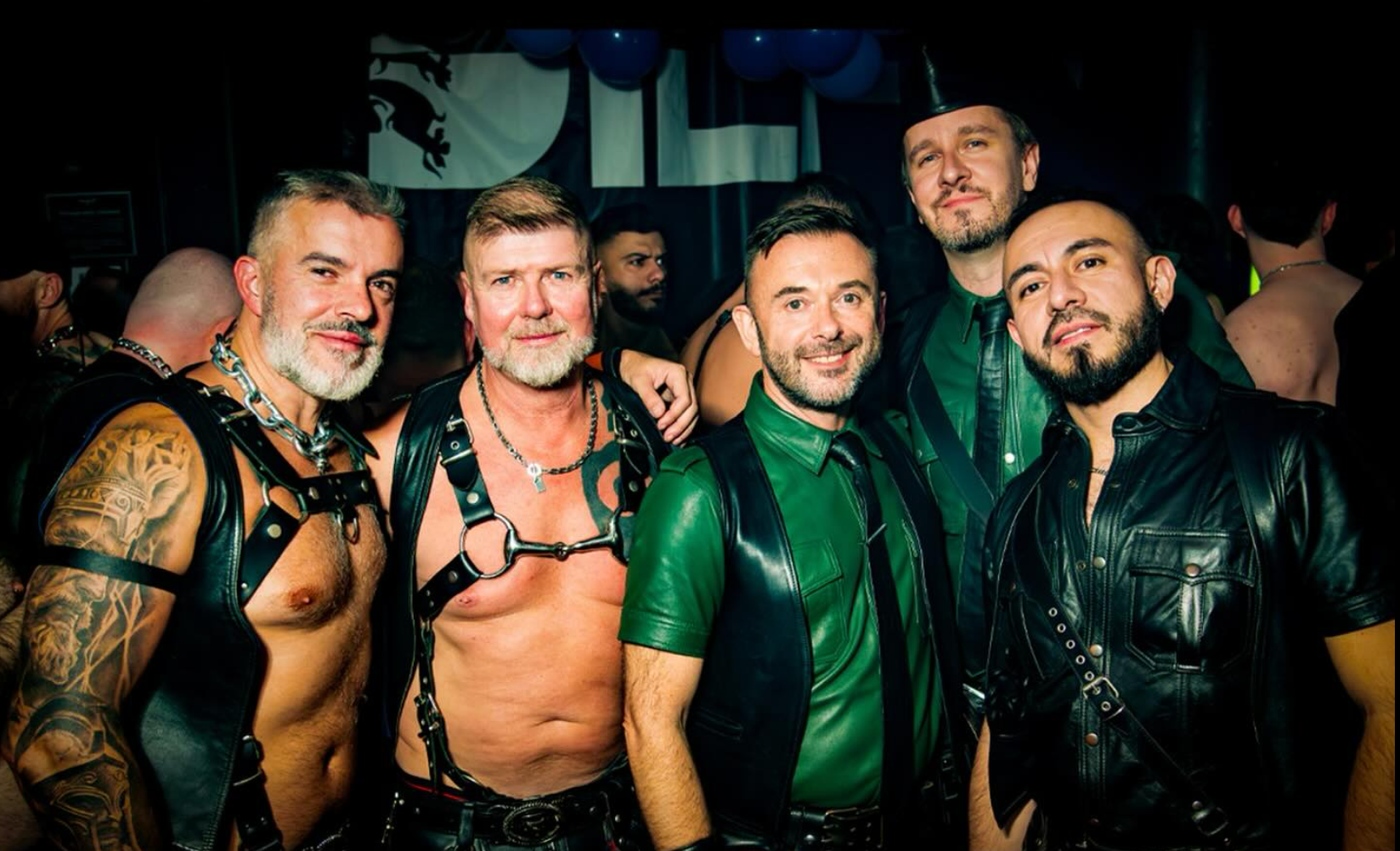 Men in leather wear at a party, some shirtless, smiling. Dark setting, various skin tones, diverse attire. Maspalomas Fetish Pride