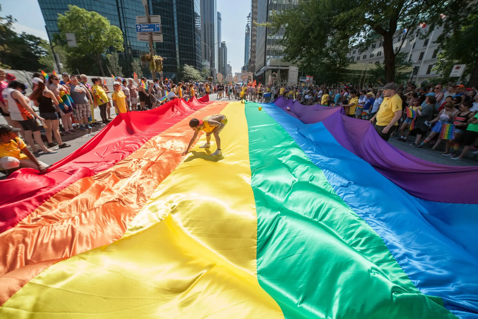 A large rainbow flag is held by participants during an outdoor parade on a sunny city street.