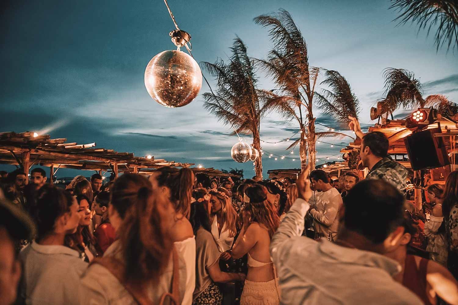 Party at outdoor venue with disco balls, palm trees, and a crowd dancing at dusk.