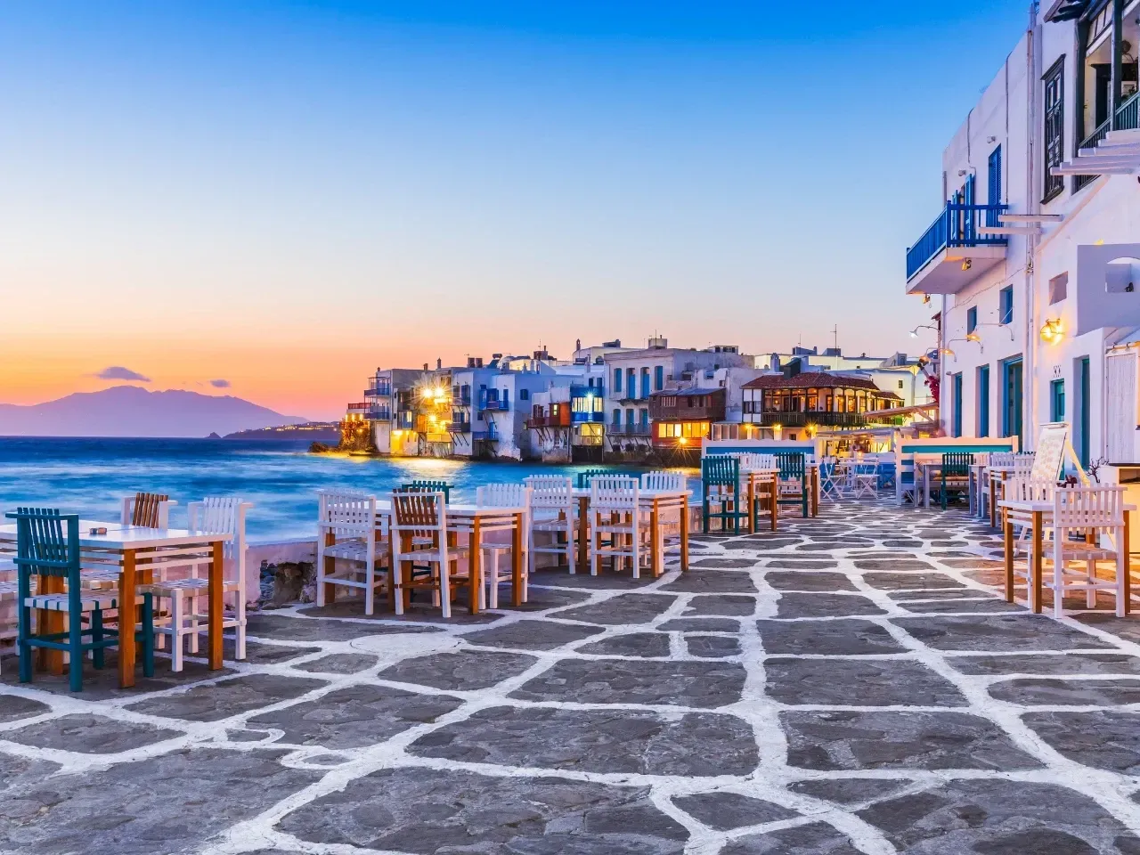Waterfront taverns with tables and chairs in Mykonos, Greece, at dusk.