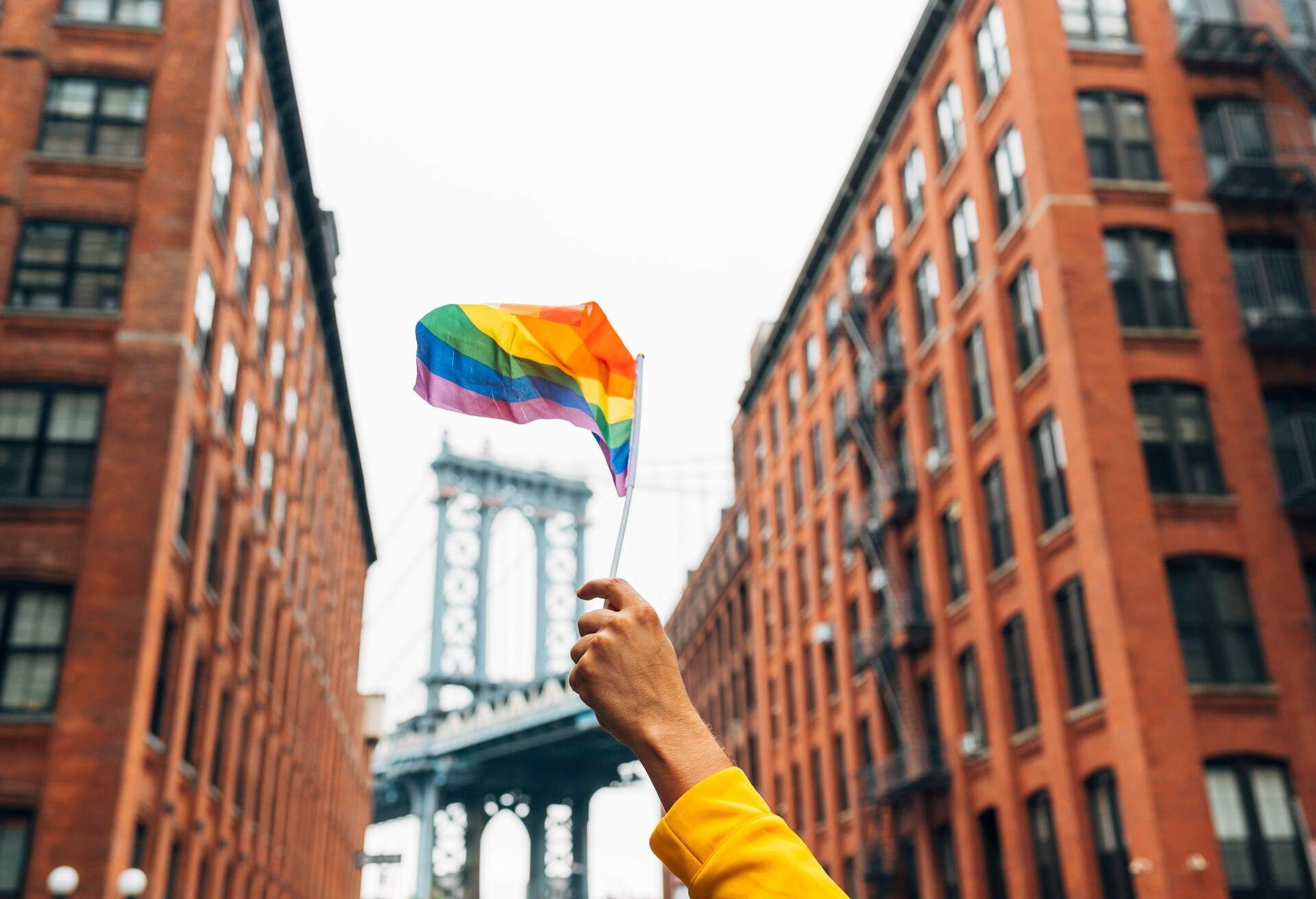 Hand holding rainbow flag in front of the Manhattan Bridge between brick buildings, celebrating LGBTQ+ pride. New York City – Birthplace of a Movement