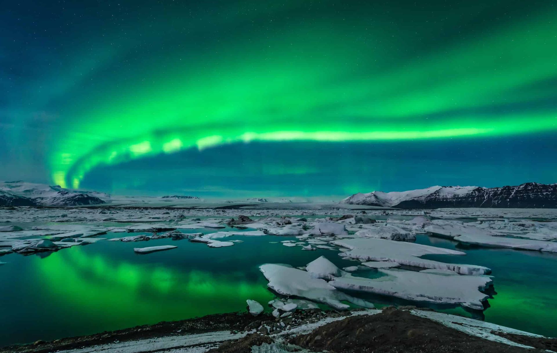 Green aurora borealis over ice-covered lagoon; mountains in the distance. Iceland