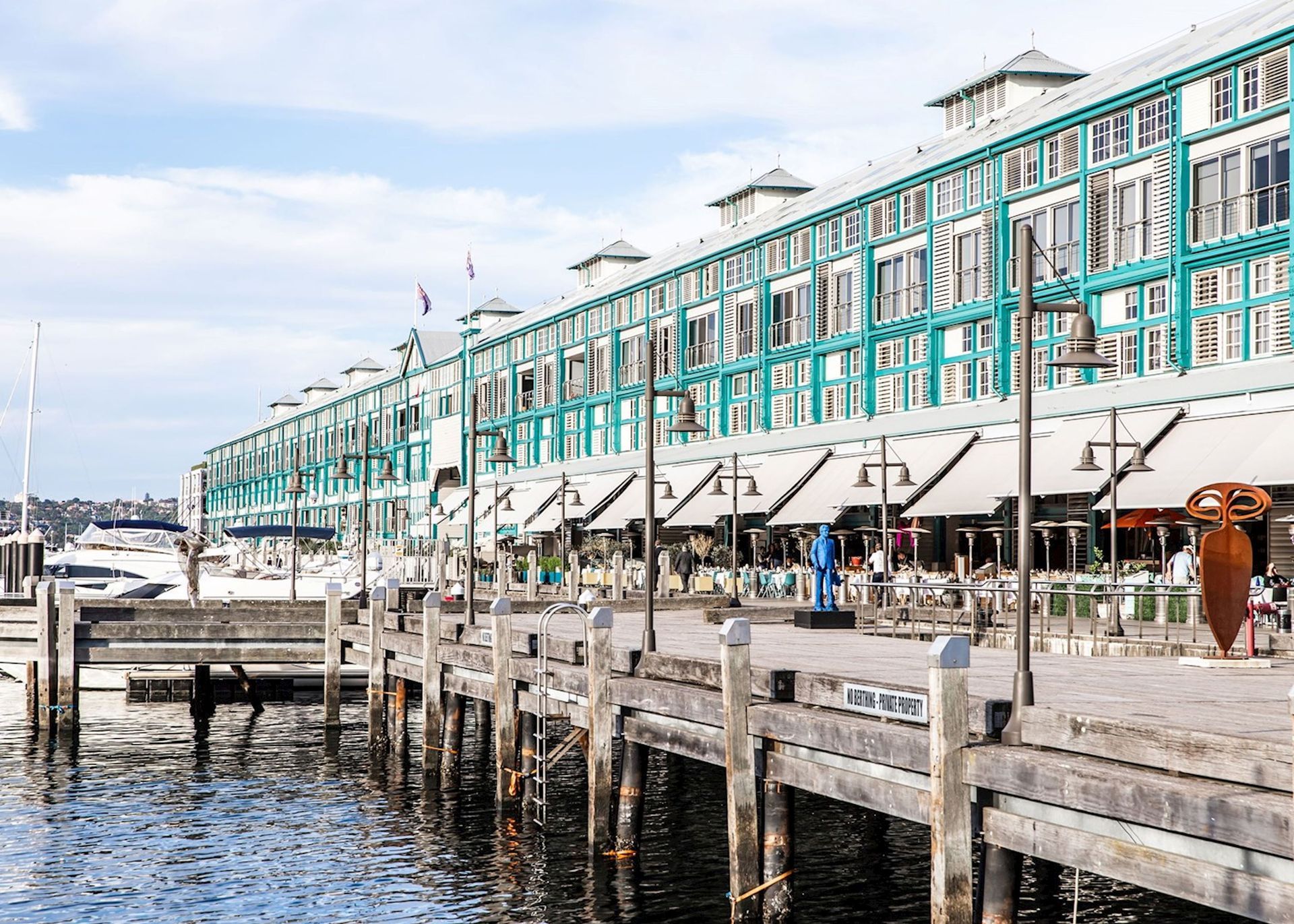 Waterfront building with teal trim and awnings, pier in foreground, clear sky. Ovolo Woolloomooloo 