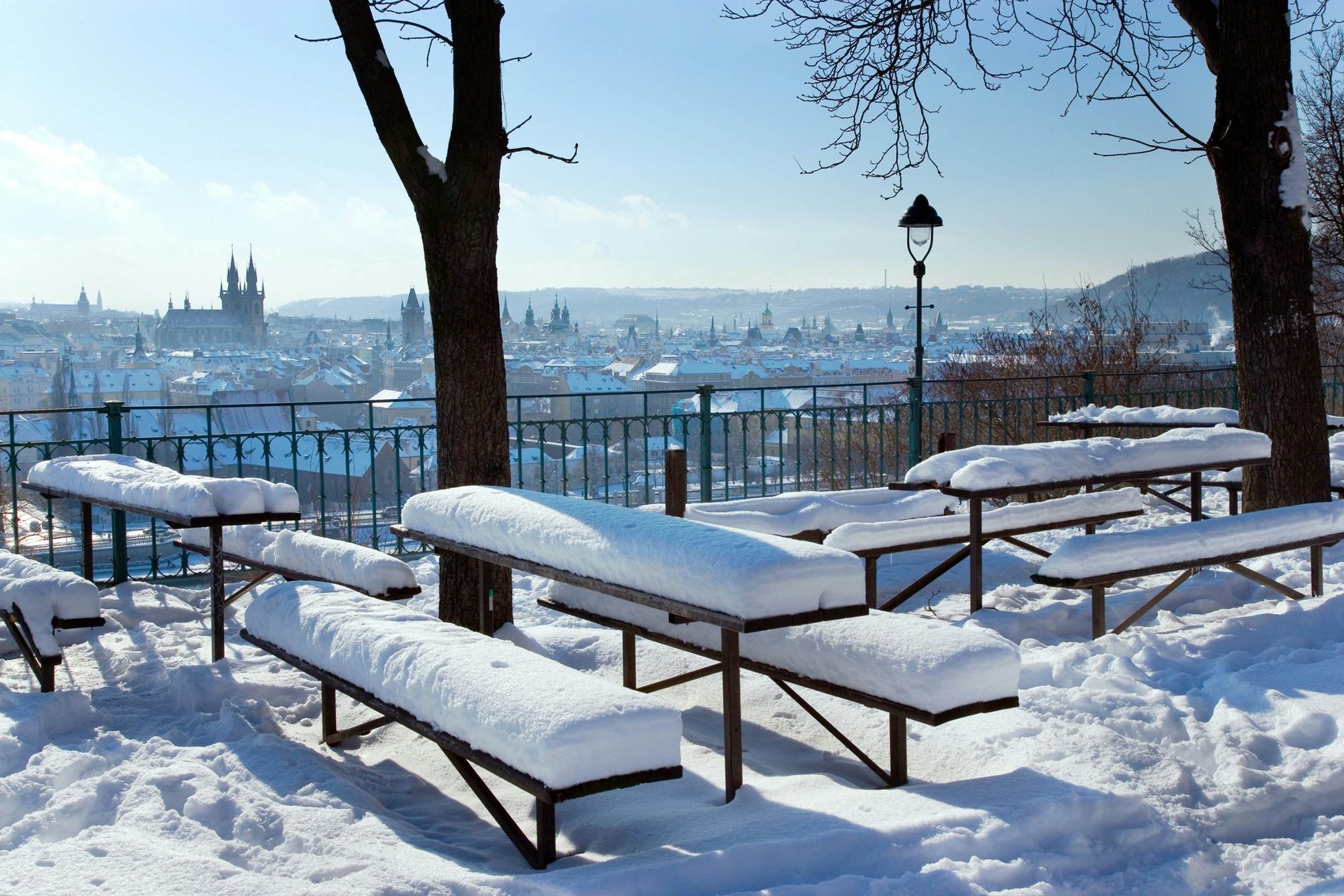 Snow-covered benches overlook a snowy cityscape on a sunny day. Prague