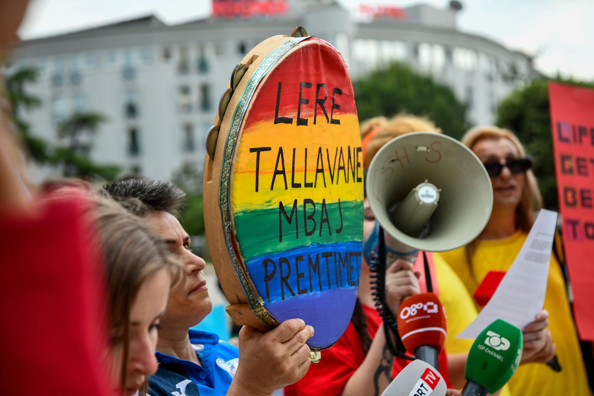 Protest with rainbow drum, megaphone, and microphones in front of a building.