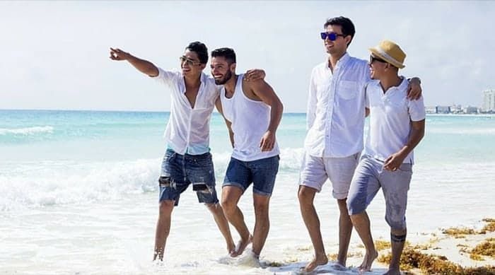 Four people walking on a beach, waves crashing. One points, they wear white shirts and shorts. Sunny day.