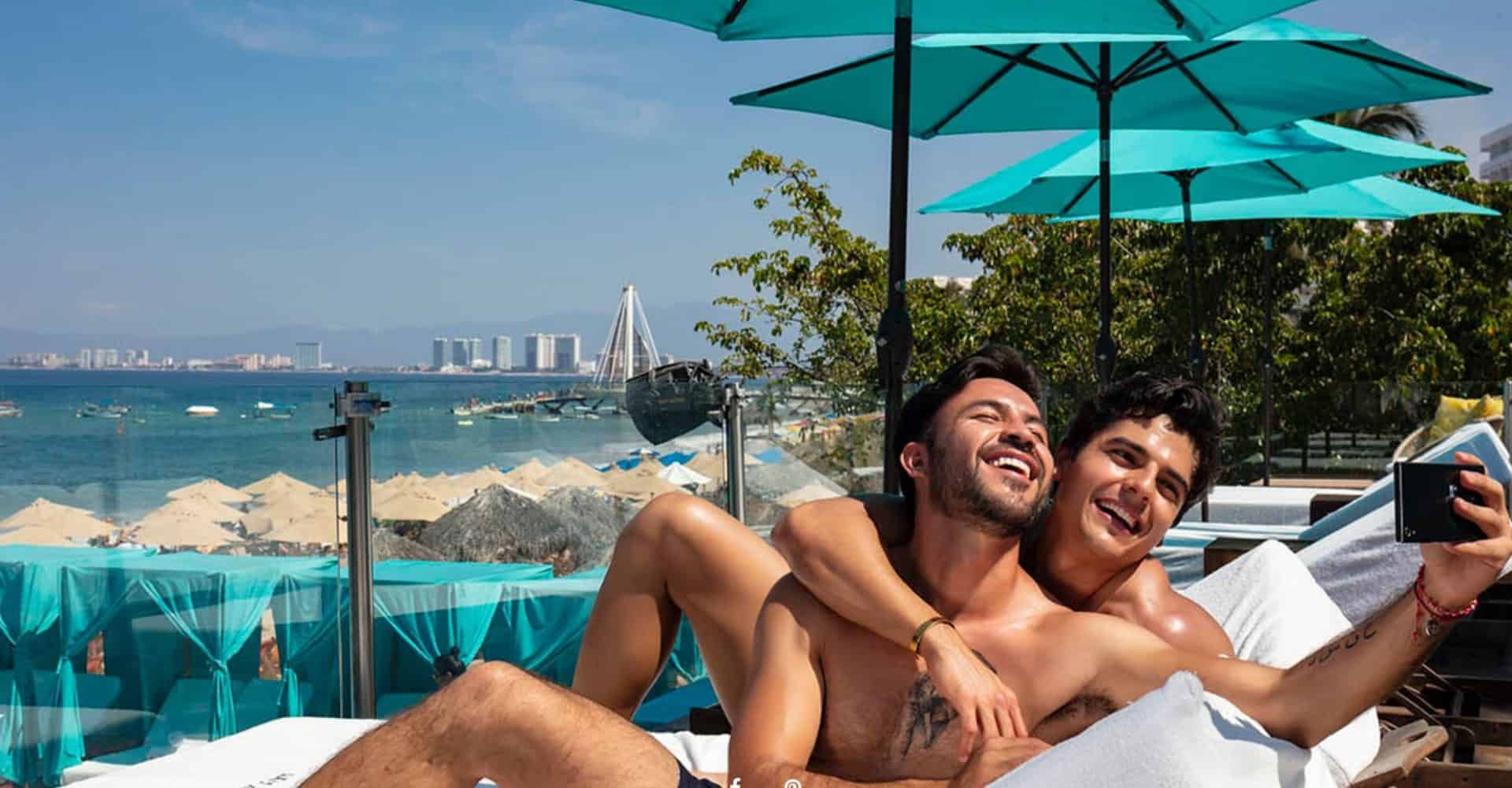 Two shirtless men, embracing, taking a selfie on a sunbed with ocean view.