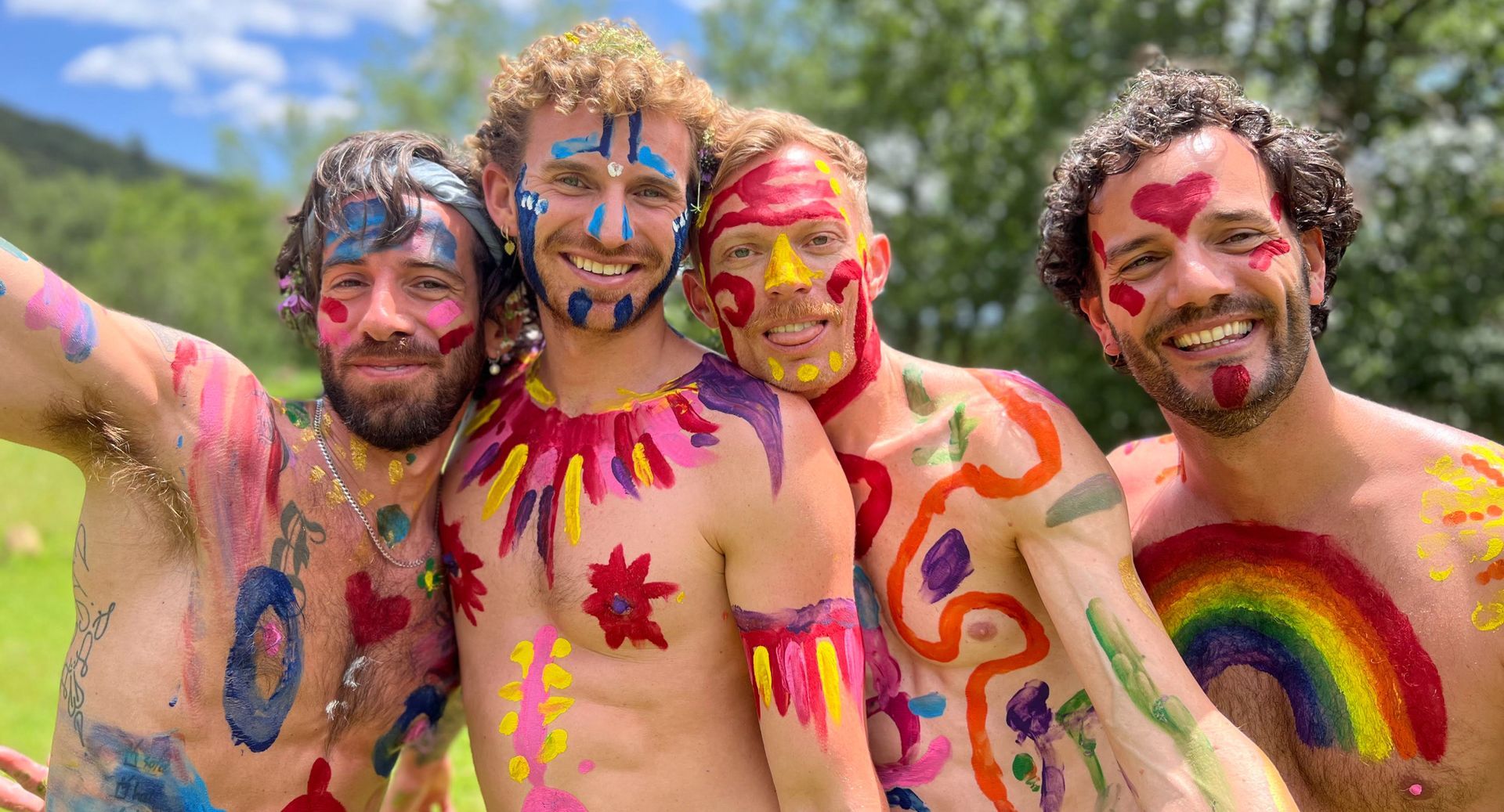 Four shirtless people with colorful body paint smiling outdoors.