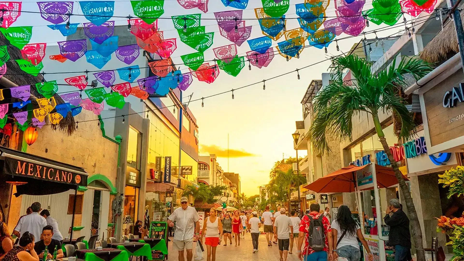 People walk along a brightly decorated street in Playa del Carmen, Mexico, with colorful flags overhead and shops. Riviera Maya