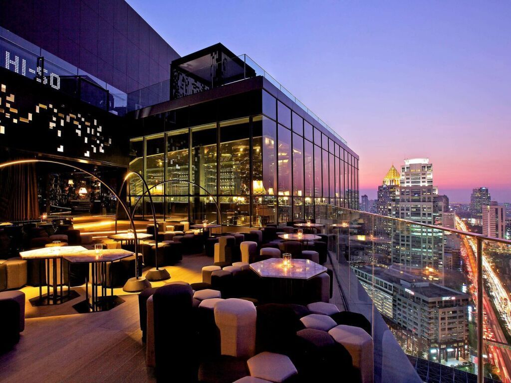 Rooftop bar at dusk overlooking a cityscape with tables, seating, and a building with the words 