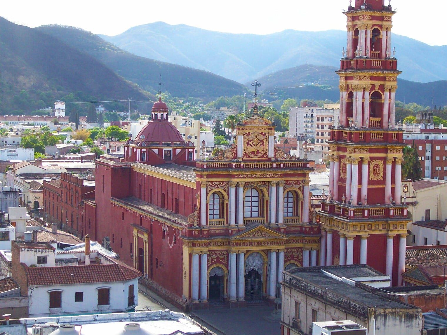 Red and gold church with a tall bell tower in a city, mountains in the background. Salta, Argentina