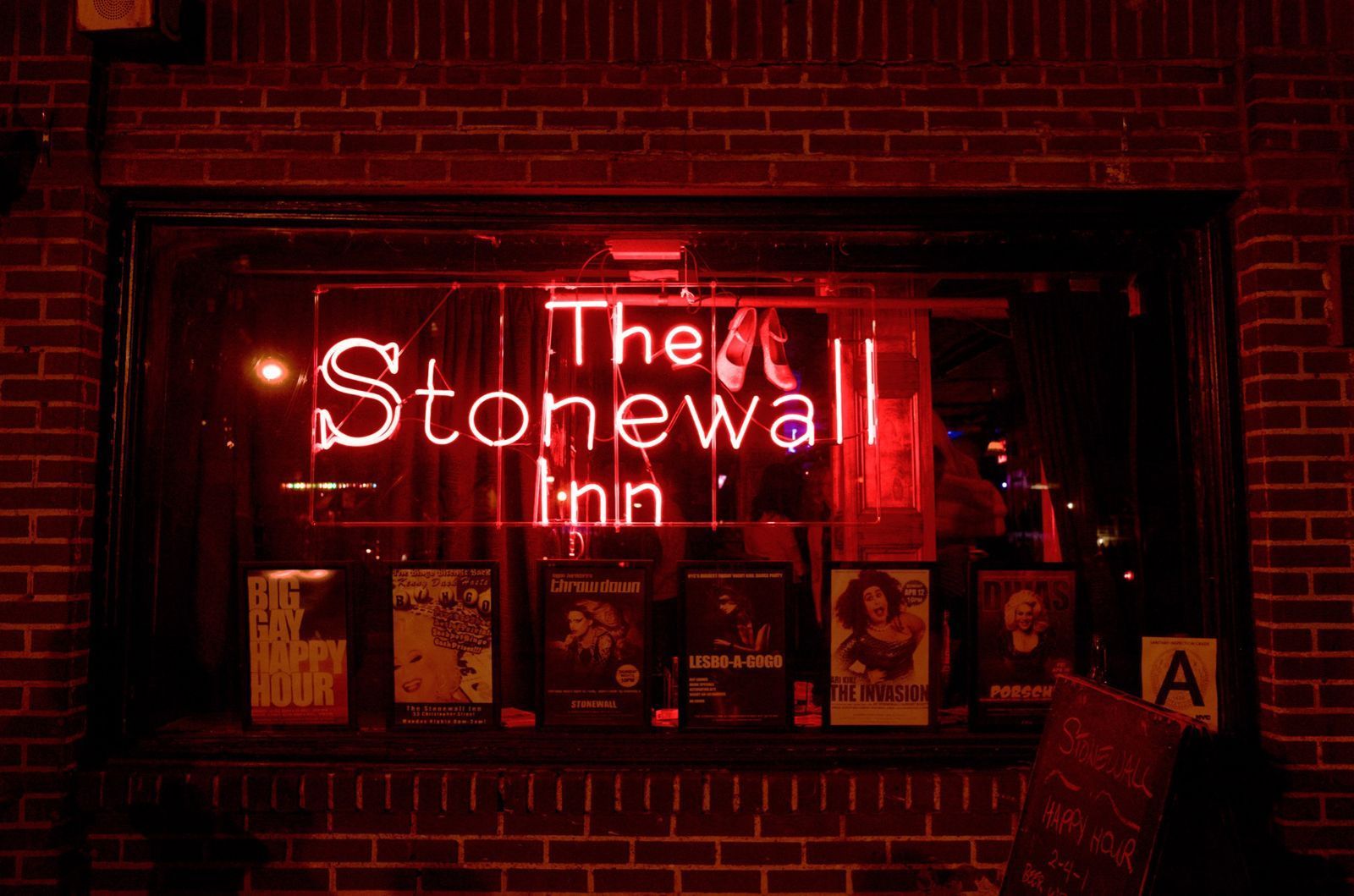 The Stonewall Inn sign in neon red, over a window display of books, set against a brick facade at night.