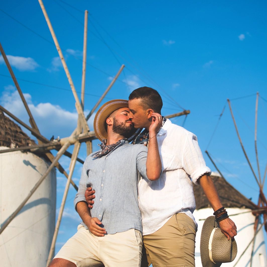 Two men kissing, arms around each other, near windmills under a blue sky.