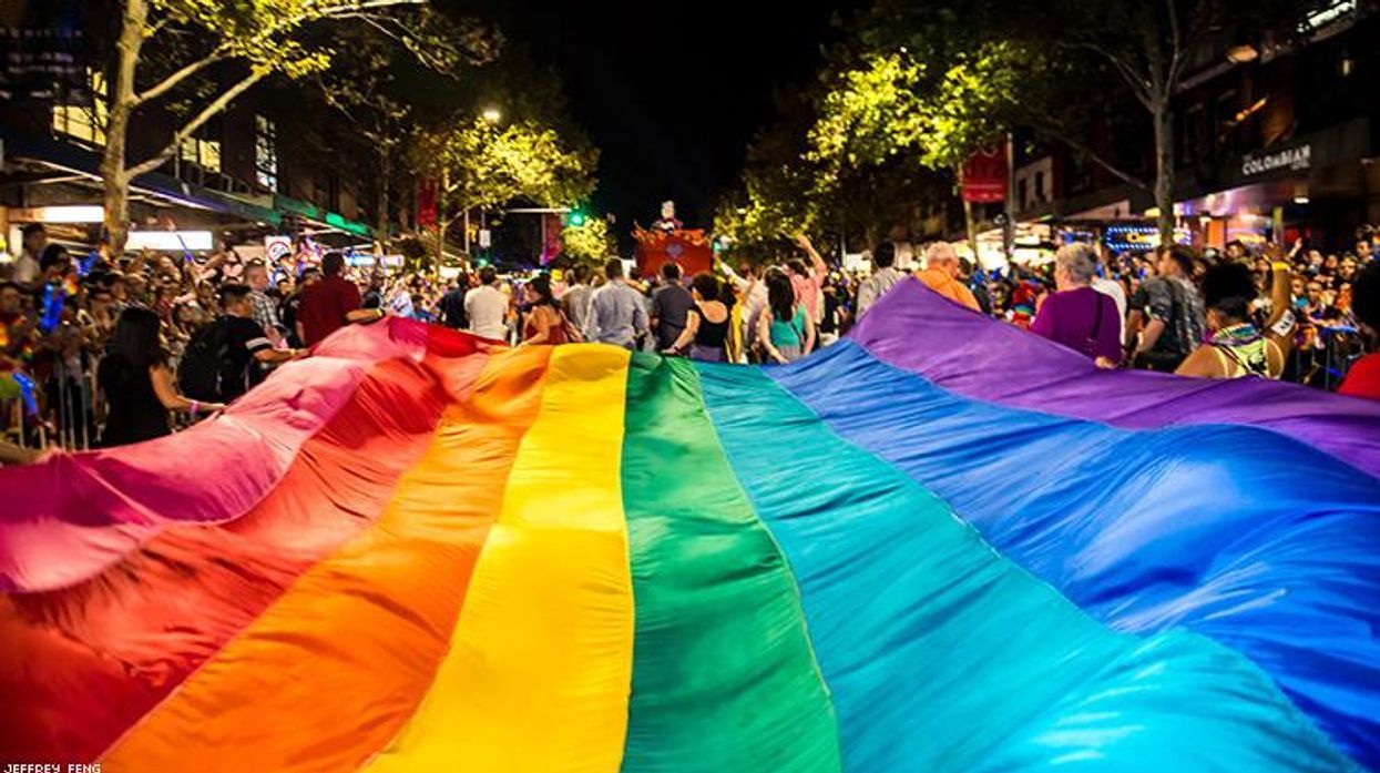 A large rainbow flag held by a crowd during an outdoor nighttime event, with colorful fabric dominating the foreground.