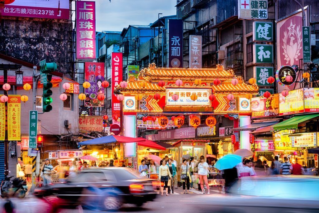 A busy city street at dusk, featuring a golden, ornate temple archway, glowing lanterns, and vertical Chinese signs.