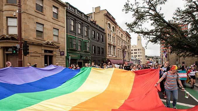 People carry a large rainbow pride flag down a city street lined with historic buildings.