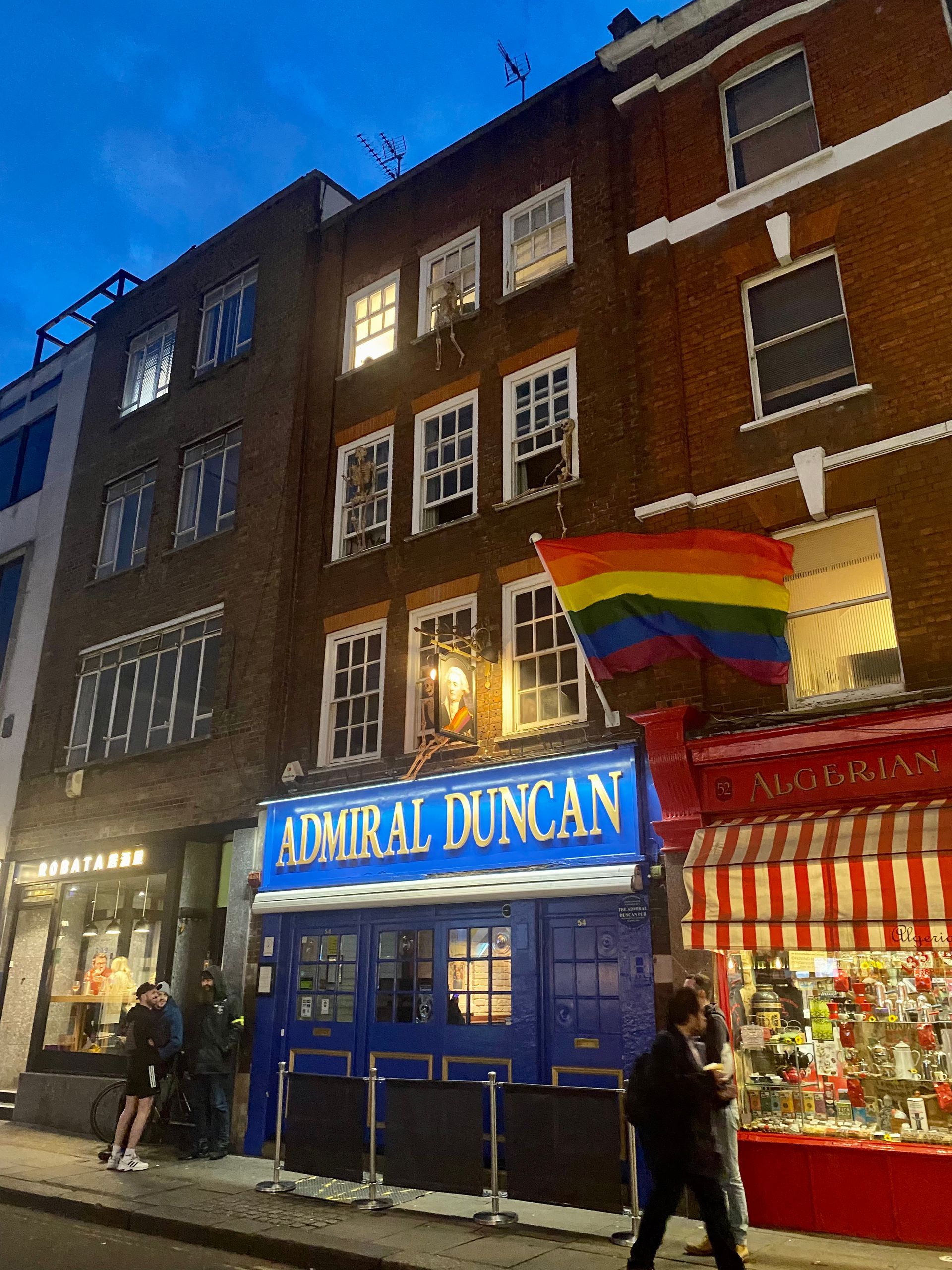 Blue pub storefront “Admiral Duncan” at night with a rainbow flag and pedestrians outside.