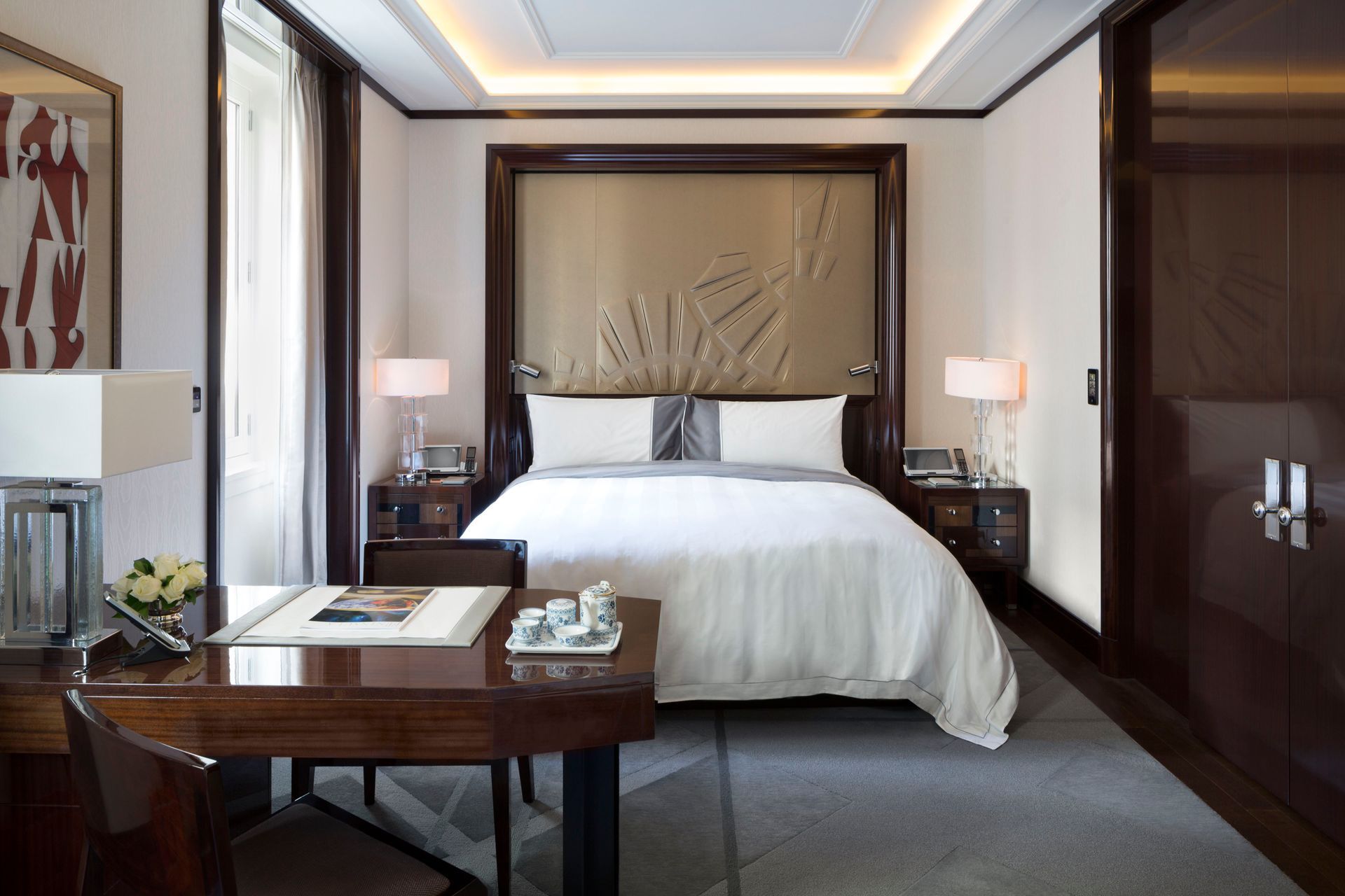 A modern hotel bedroom featuring a queen bed with white bedding, a desk in the foreground, and ambient warm lighting.