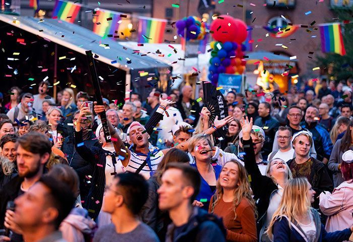 A cheering, diverse crowd celebrating at an outdoor Pride festival with rainbow flags and confetti filling the air.