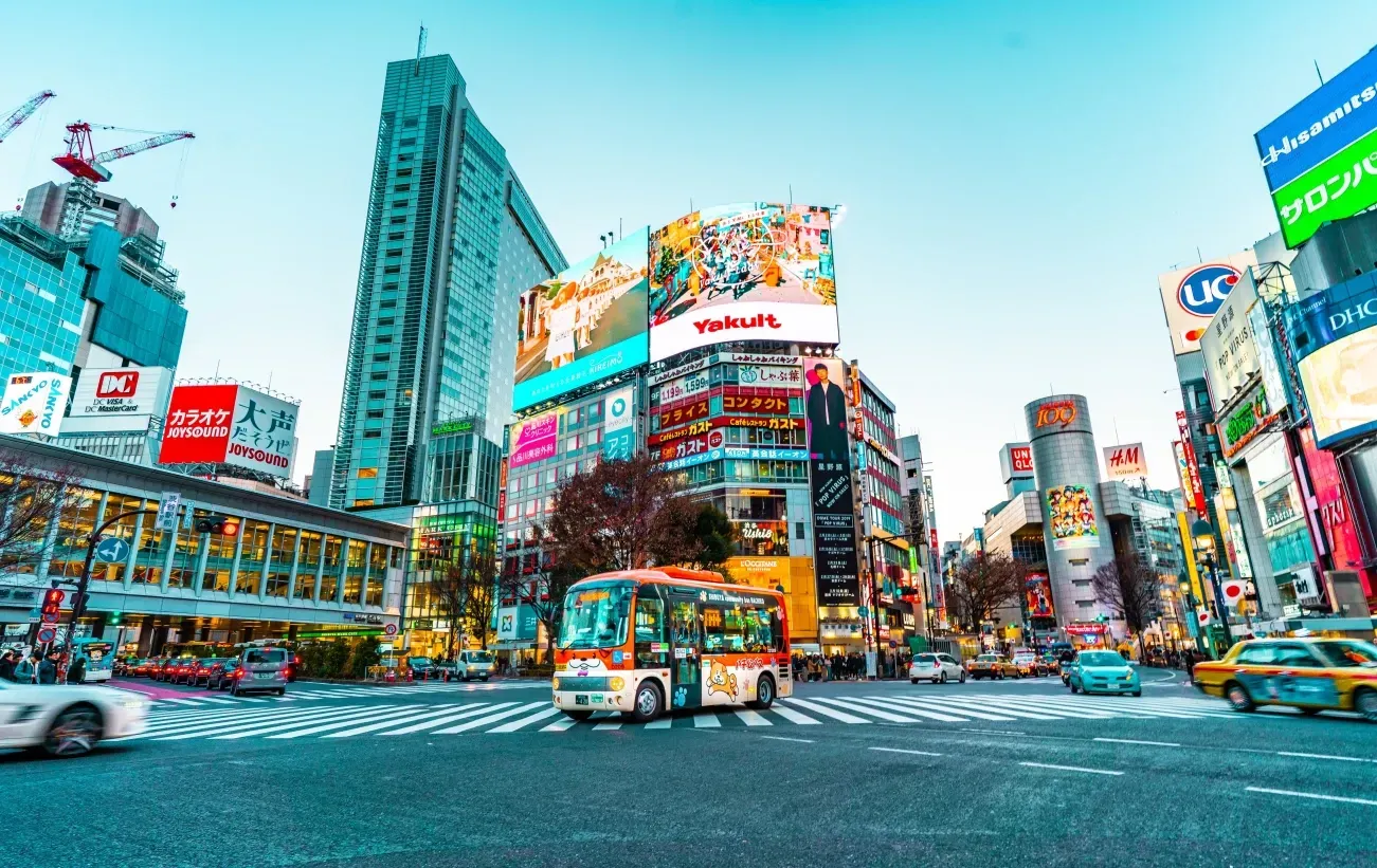 A bus travels through the busy, neon-lit Shibuya Crossing in Tokyo, surrounded by towering buildings and advertisements.