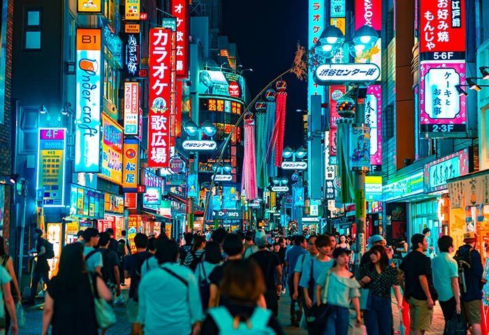 Crowd of people walking through a brightly lit street in Japan, lined with neon signs. Tokyo