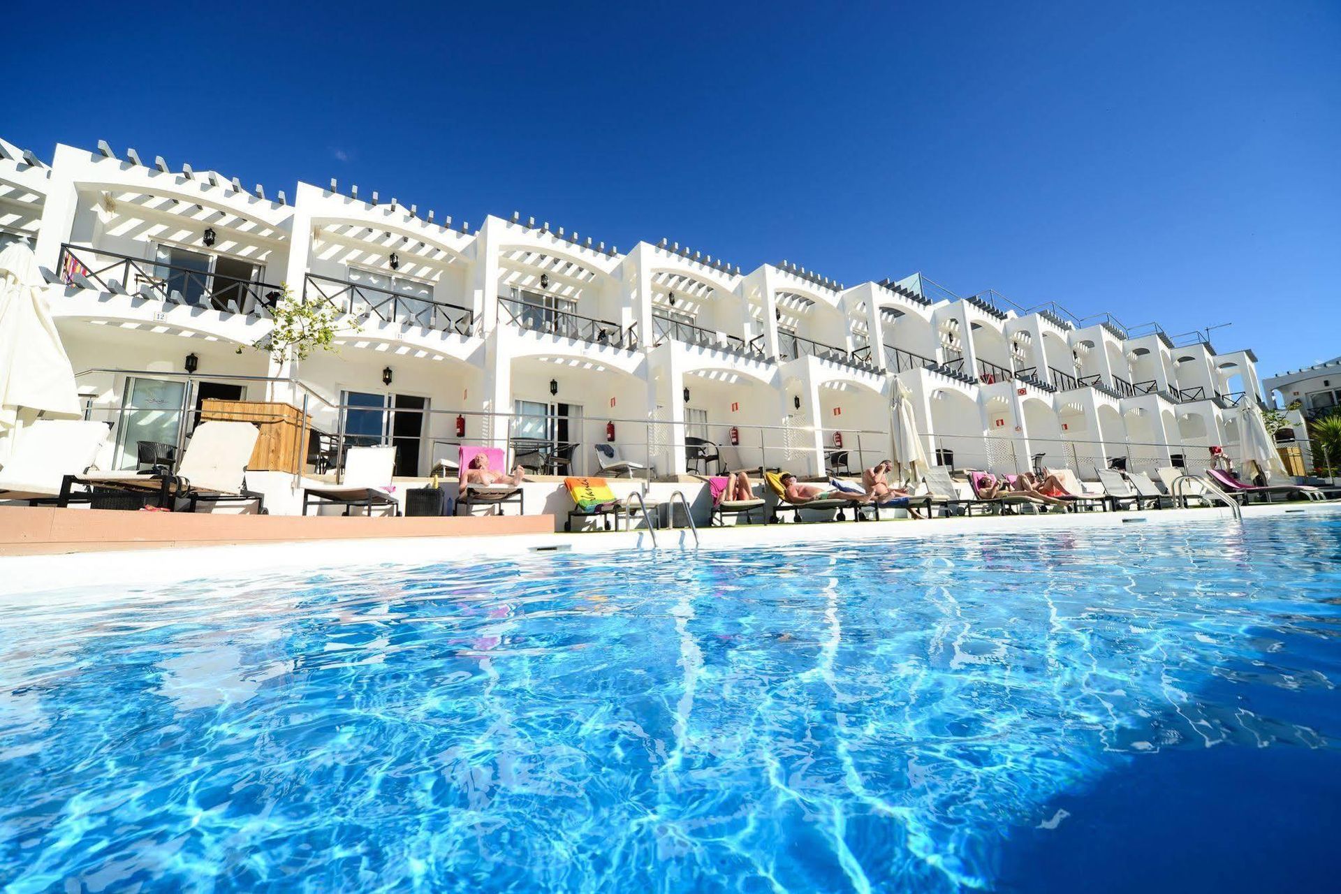 Pool in front of a white resort building with balconies under a bright blue sky.