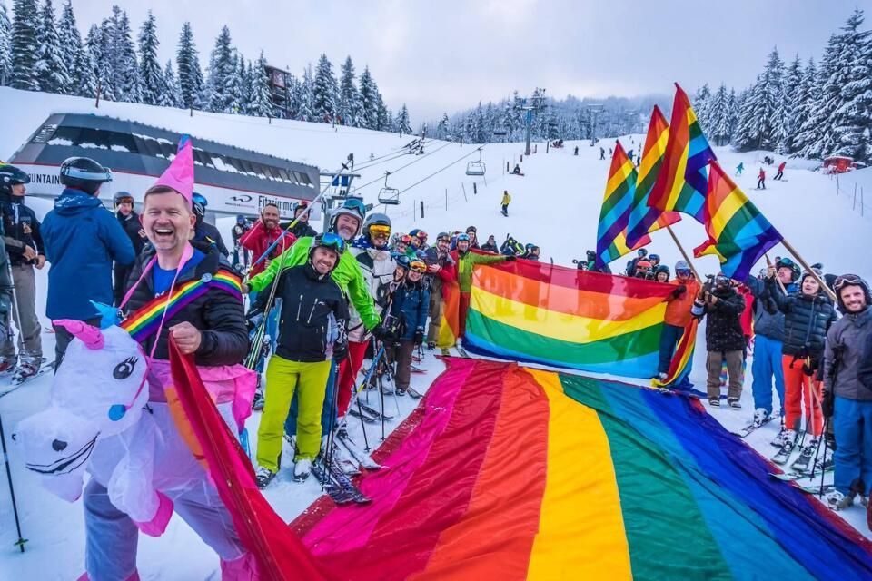 Group of skiers celebrating with rainbow flags and attire on a snowy ski slope. Whistler Pride