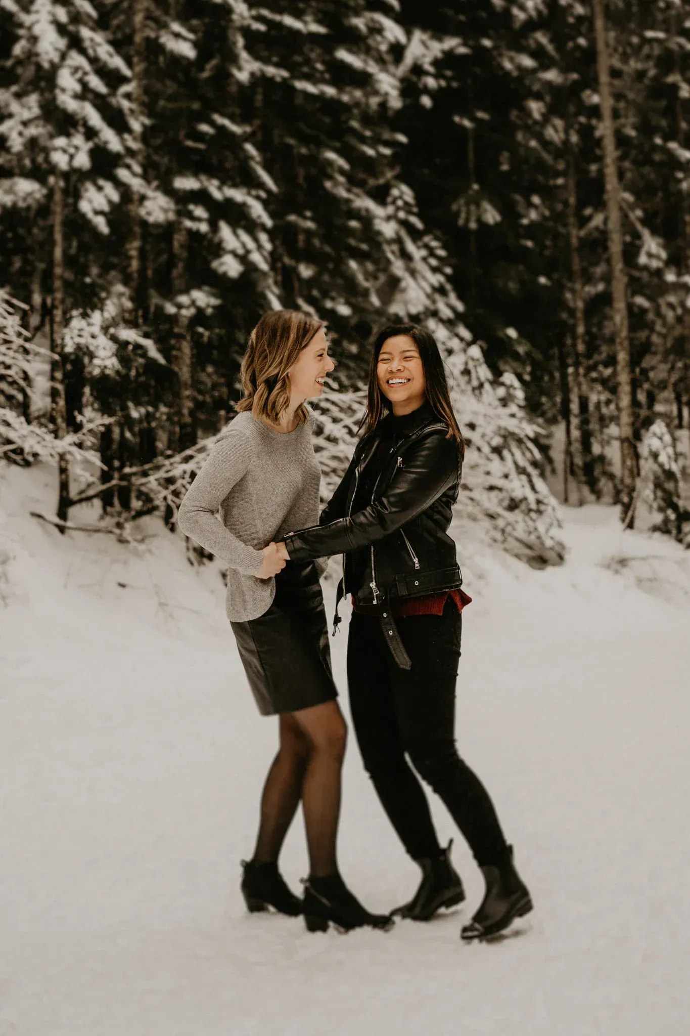 Two people hold hands in a snowy forest. One wears a sweater and skirt, the other a jacket and pants.