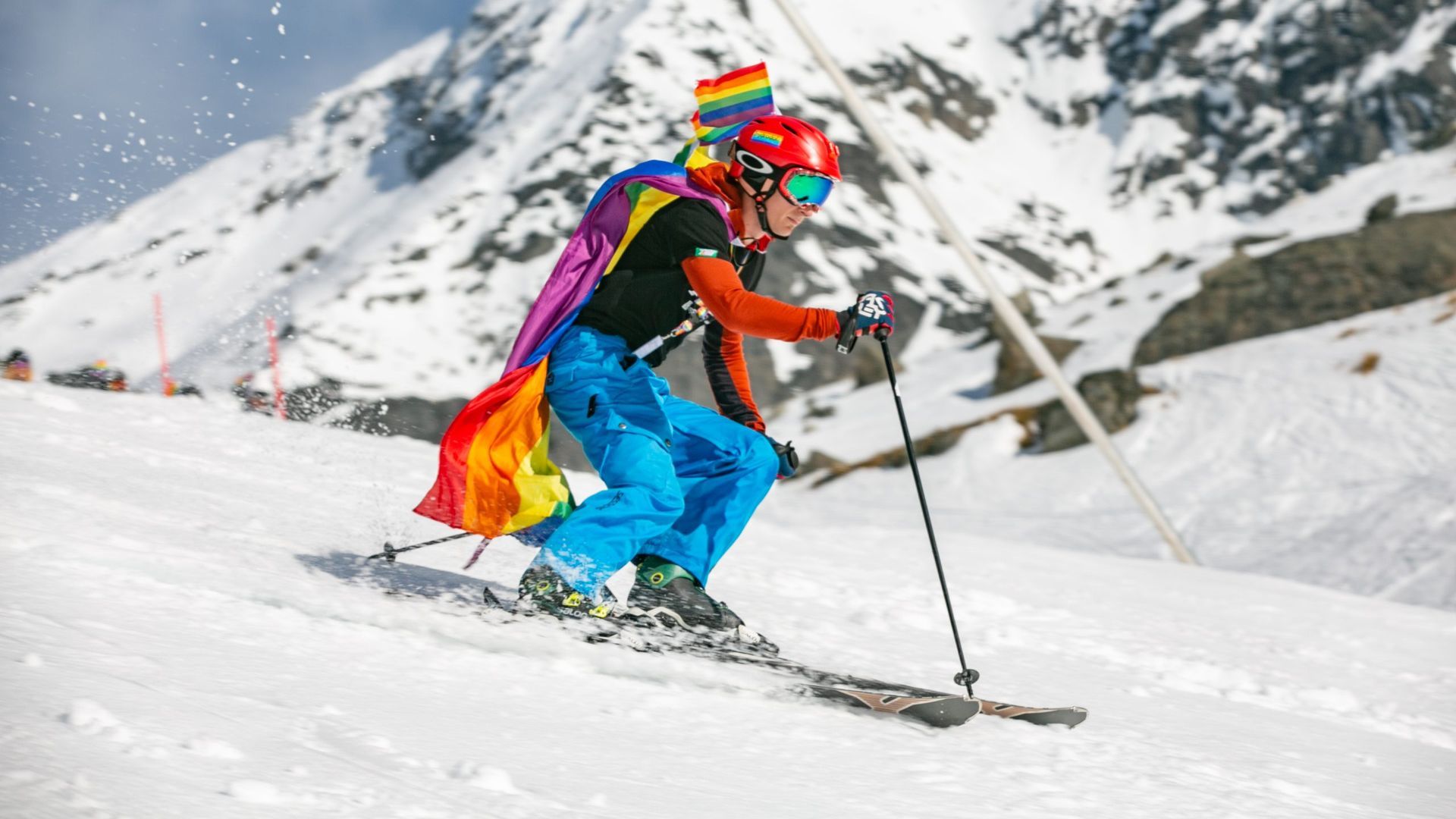 Skier with a rainbow flag cape on a snowy mountain slope.
