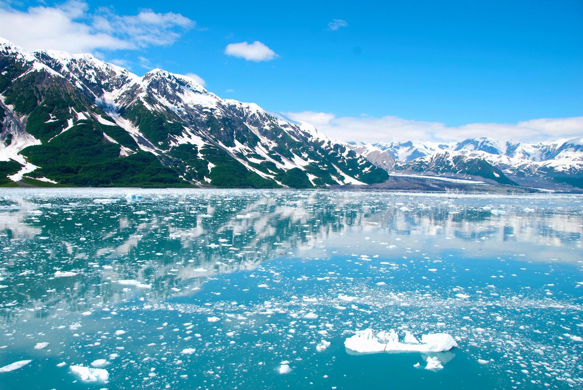 Snow-capped mountains reflecting in a turquoise ocean filled with ice, under a bright blue sky.
